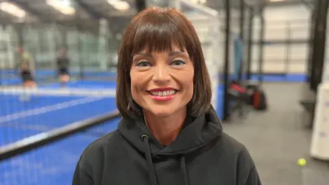 Sam Read/BBC A woman with shoulder length brown hair and a fringe stands in a black hoodie while padel is played in the background.