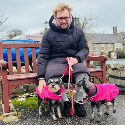 Picture of a man with curly hair blowing in the wind, sitting on a bench, with two dogs in pink jackets at his feet
