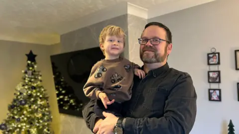 John holds Ralphie in his arms. They are standing in a living room, with a Christmas tree behind them.