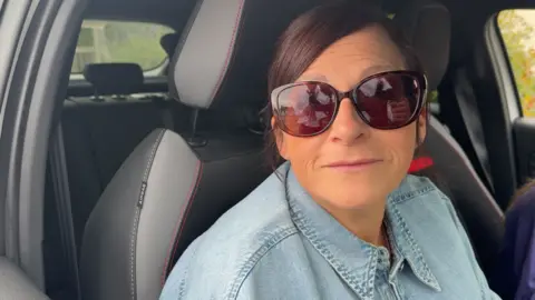 A woman in a denim shirt with burgundy hair (and sunglasses to match) is sitting in her car looking out of an opened window.