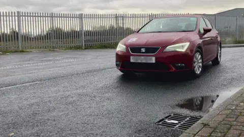 Seb Cheer/BBC A red hatchback car driving along a street with an L plate on the bonnet.