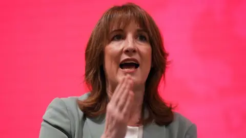 Rachel Reeves speaking in front of a red backdrop. She has shoulder length brown hair, her right hand is raised and she's wearing a light green coloured suit.