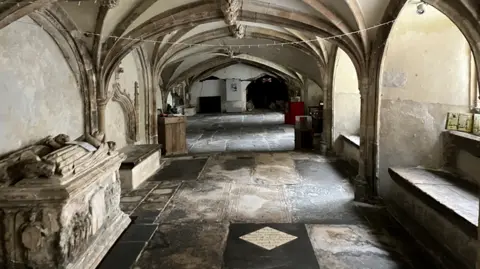 The inside of the crypt. Vaulted cielings can be seen and there are tombs on the left side of the stone crypt. Different coloured flagstones can be seen on the floor.