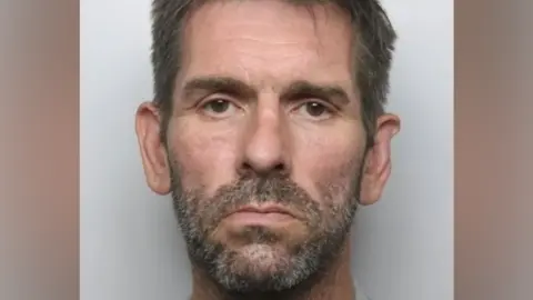 A man with dark hair and grey speckled facial hair stares at the camera. He is not smiling