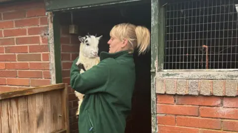 Jamie Dowling, wearing green work clothes, holds a goat in her arms