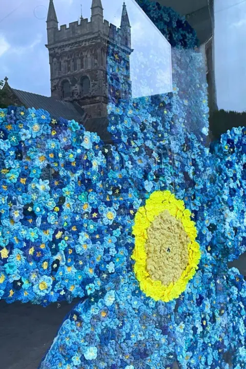 Museum of East Dorset A close image of the blue knitted flowers in the window of the museum with the reflection of the Wimborne Minster in the window