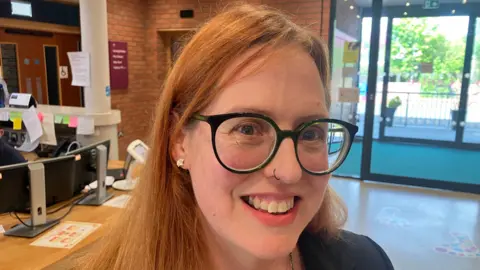 A woman with auburn hair smiles at the camera. She is wearing glasses and is standing next to a wooden desk with computer monitors on it. 