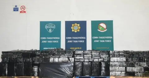 AN GARDA SÍOCHÁNA Piles of packages wrapped in black plastic in front of three signs saying 'joint task force' 