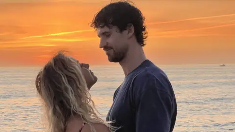 Matthew MacNabb Matthew and Laura looking at each other. They are standing in front of the sea and the sunset is bright orange. Laura has long blonde hair. Matthew has black hair.