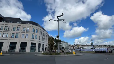 A tall silver column, with the three legs of mann, stands above a roundabout. There is a modern light building in the background and marquis tents on the other side.