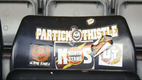 Patrick Thistle Ultras stickers on a seat in Firhill Stadium. The stickers have black, red, yellow and white logos and text.