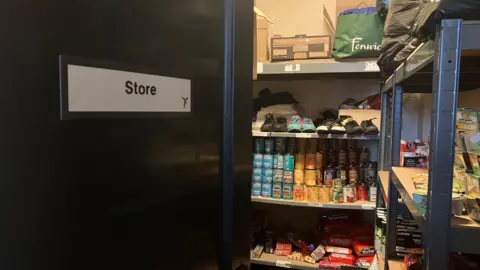 The inside of a small storage room labelled “Store” on a sign attached to the open door. Inside, metal shelving units are filled with a variety of items, including: Canned goods (beans, soups, vegetables) stacked in rows
Food items such as biscuits, snacks, and packaged products
Clothing items, including several pairs of shoes on an upper shelf