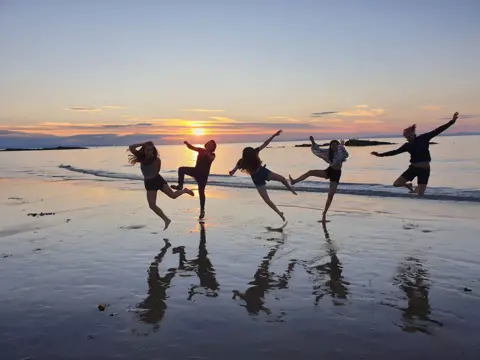 Jacky Pratt A group of people dance and jump on a beach as the sun sets