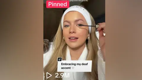 TashaAmberGhouri/TikTok TikTok video showing Tasha, a white woman, holding a black mascara wand, while applying it on her left eyelash. She has a white headband on and is wearing a white gown. The title of the video written in a white box and black font is "embracing my deaf accent" white a white heart emoji. Under that is a white "play" sign with 2.9m written. At the top of the screenshot is a "pinned" caption written in white on a pink box.