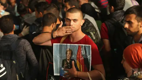 Getty Images A migrant from Syria holds a picture of German Chancellor Angela Merkel as he and approximately 800 others arrive from Hungary at Munich Hauptbahnhof main railway station on September 5, 2015