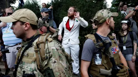 Reuters man in white suit standing behind two heavily armed men in bulletproof vests