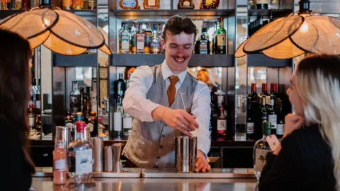 BBC A man pours a cocktail while two women watch at Ricks Bar at the Bristol Hotel