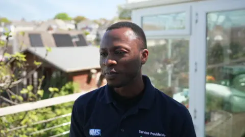 A man called Abiodun Olafusi. He is black and is wearing a navy blue polo shirt with the NHS logo on it. He is stood next to a conservatory at a home.