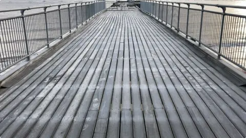 Luke Deal/BBC The length of the Claremont Pier. The end of it has been gated off to the public. Wooden boards line the surface of pier's floor. 