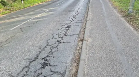 Vikki Irwin/BBC A single carriageway road and pavement. on one side of the road you can see cracks forming in the road surface which runs all the way along the road.
