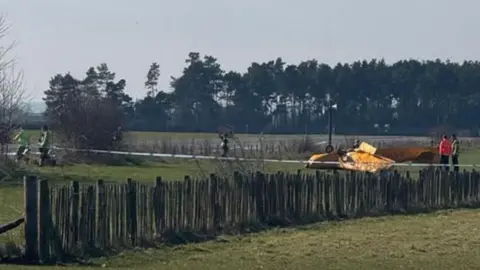 An upside down light aircraft, yellow or orange in colour. People are stood around the crashed aircraft, with a police cordon stretching across the image. A small fence also runs across the picture, with trees in the background. 