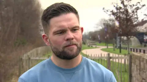 Boxer Conrad Cummings standing in a park talking to BBC News NI.  He  has short dark hair and a beard.  He is wearing a blue crew-neck sweatshirt.