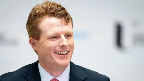 PA Media Joe Kennedy smiles. He has ginger hair and freckles. He is wearing a white shirt with a red tie and a blue blazer.  The background behind him is blurred.