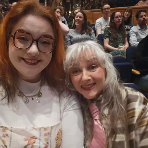 Anita West Two women sitting in the seats at a concert - one woman has long red hair and a white top on. The other has blonde/silver hair and a patterned top in shades of beige and brown