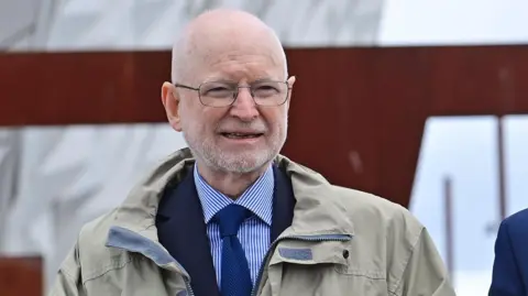 Sir Desmond Rea, a man with a shaved head and a short, white beard, walks towards the camera outside the Titanic Belfast building in 2022. He is wearing glasses, a grey/beige anorack over a navy suit jacket, blue and white shirt and a navy tie. 