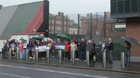 A group of about 30 pro-Palestinian protesters gathered outside Musgrave Street PSNI station in Belfast city centre. Some are holding banners and umbrellas. A large Palestinian flag is being held up in the middle of the group.