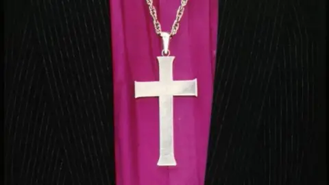 A cross on a purple garment, there is black clothing on either side.