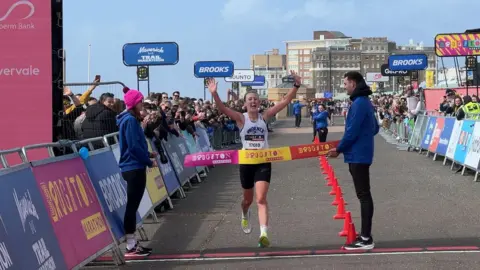 A woman in a white running top crosses a red line, breaking through a pink and yellow tape with her hands in the air. Dozens of people watch on from behind sponsor boards either side of the road.