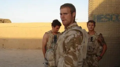 Three people including Mark Omrod in desert camouflage gear standing on sandy ground near a wall and cylindrical structure outdoors.