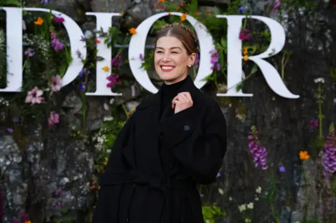 PA Rosamund Pike wearing a slouchy belted trench coat over a black turtleneck at the Dior catwalk