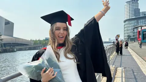 Ella is standing in her graduation gowns next to Salford Quays. She is holding flowers in one hand and waving her other in the air. She has a joyful smile. 