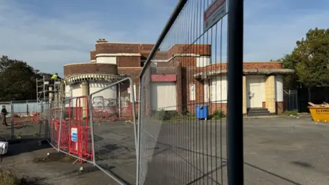 Andrew Turner/BBC The Iron Duke, with fencing in the foreground. The building is two storeys with many rounded features, terracotta mouldings and curved windows, which are boarded up.