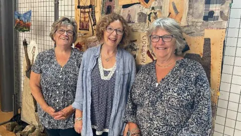 A photo which shows three women stood next to each other. The woman on the left has short blonde-ish hair and glasses, she is wearing a black and white patterned top with white buttons up the front, she is holding her hands together in front of her. The woman in the middle has shoulder length hair and is wearing glasses, she is wearing a blue long jacket and a blue and white patterned top. The woman on the left has short hair, glasses and a blue and white top.