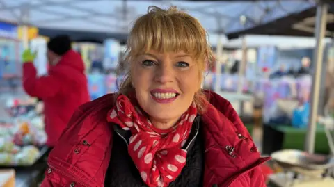 A woman with blonde hair, wearing a red coat, black top and red-and-white scarf. She is standing behind a market stall under a canopy, and smiling at the camera. 