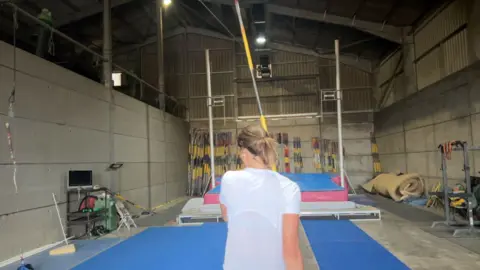 Gemma Tutton, wearing a white athletics vest and carrying a pole, runs towards a vaulting bar.