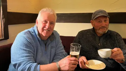 Two men smile at the camera one is holding a pint of beer, the other holds a cup of tea, they are sitting in a pub. 