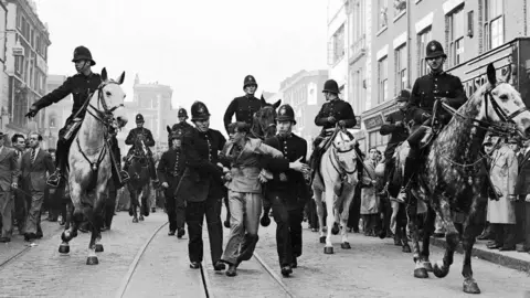 Alamy A black and white image of an  anti-fascist demonstrator being taken away under arrest by police after a mounted baton charge during the Battle of Cable Street, London, 4 October 1936 