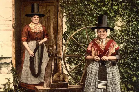 Universal Images Group via Getty Images Two women stand next to a large wooden spinning wheel. They wear traditional Welsh clothing - a black chimney hat, red shawls, and black and white aprons. 