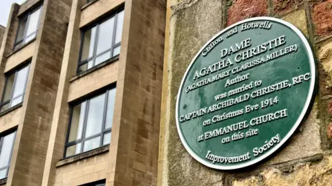 A large green plaque on the side of a stone wall in Bristol explains how the author Agatha Christie was married there in 2014. In the background is a more modern building with dark frames and stylish light brown bricks