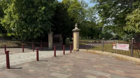 The stone and metal entrance gates of Barnes Park. A banner promoting an upcoming family fun day is hanging on the red fence.