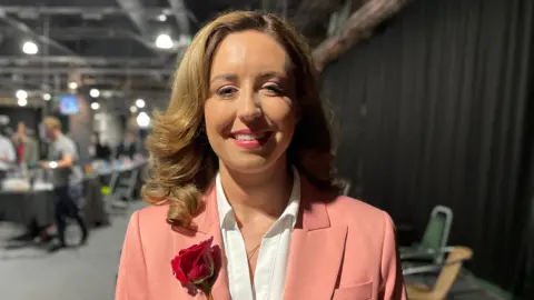 A woman has light brown shoulder length hair which is slightly curly. She is wearing a white shirt and a peach blazer with a red rose pinned to the lapel. She is smiling at the camera