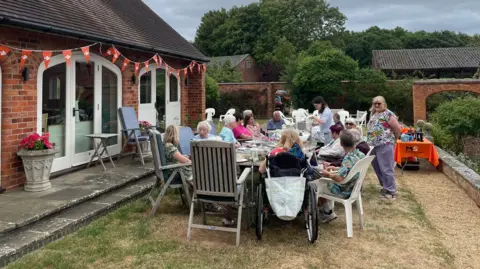 Jacqueline Krarup A group of people sitting around a garden table. There is bunting for decoration and an MS Society branded table in the corner. One woman in a floral shirt is standing smiling at the camera, while the others are seen socialising with each other.
