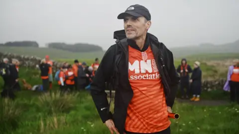 Andrew Hartley dressed in a orange t-shirt with the words MND Association stands in a field near a stretch of Hadrian's Wall in Northumberland.
