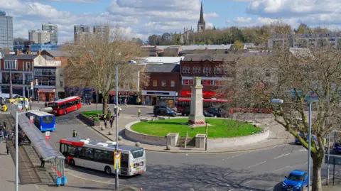 An image of Walsall on a sunny day. We can see a war monument on a roundabout and a number of buses at a nearby bus stop. There are trees and there are not many people about. There are a few cars going around the roundabout. 