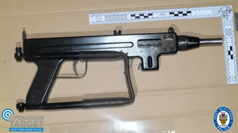 A black gun with a big handle and large trigger on a brown table with a metal ruler lying alongside it to show its size