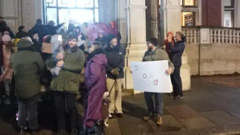 Rebecca Brueton A group of protesters gathered outside. One is holding a sign which says 'save our pools'.